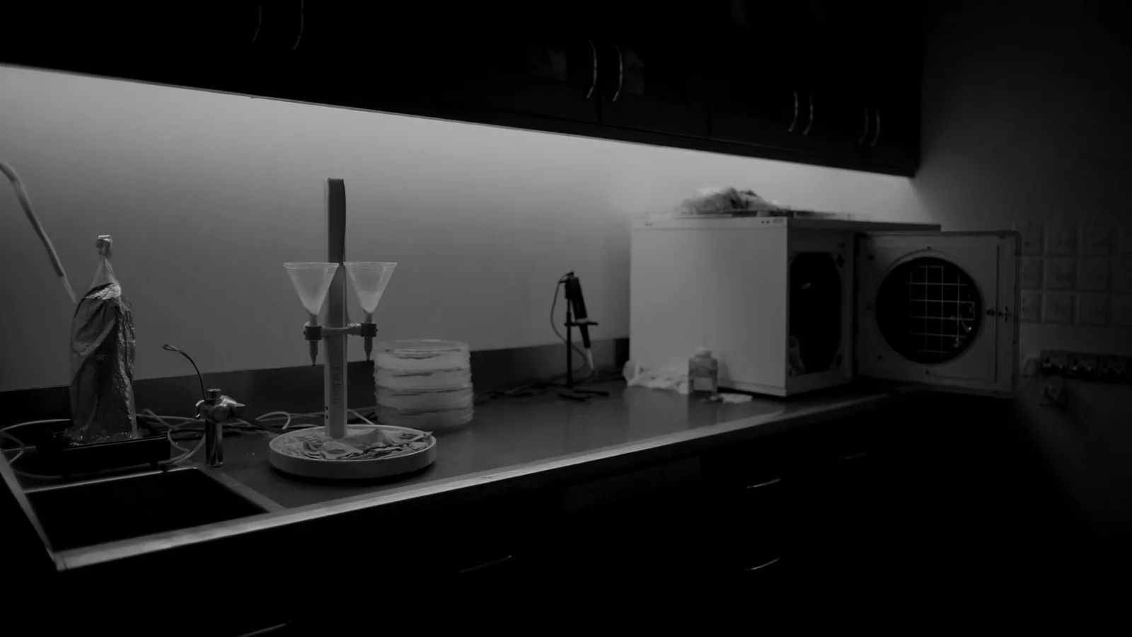 A laboratory bench with a membrane filtration manifold, stacked petri dishes and a pipettor, under fluorescent light.