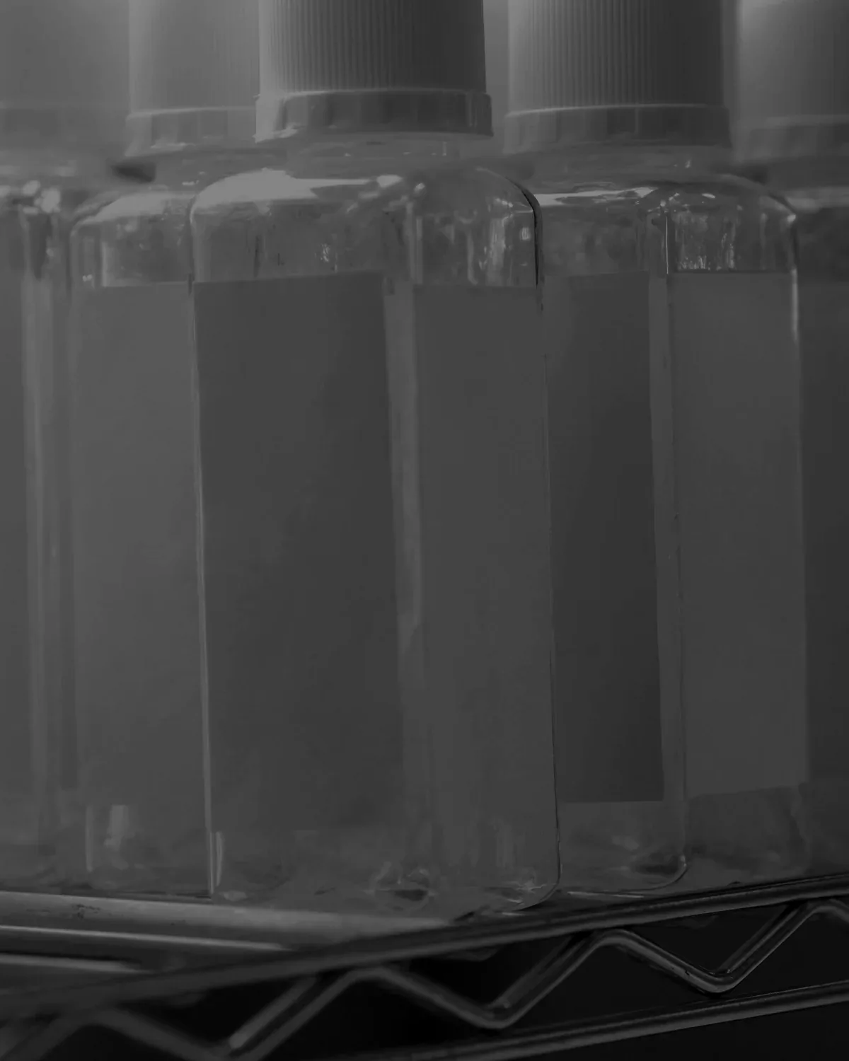 A wire rack of sterile plastic sample bottles with white screw caps and blank labels, condensation on the outside, on a stainless steel counter.
