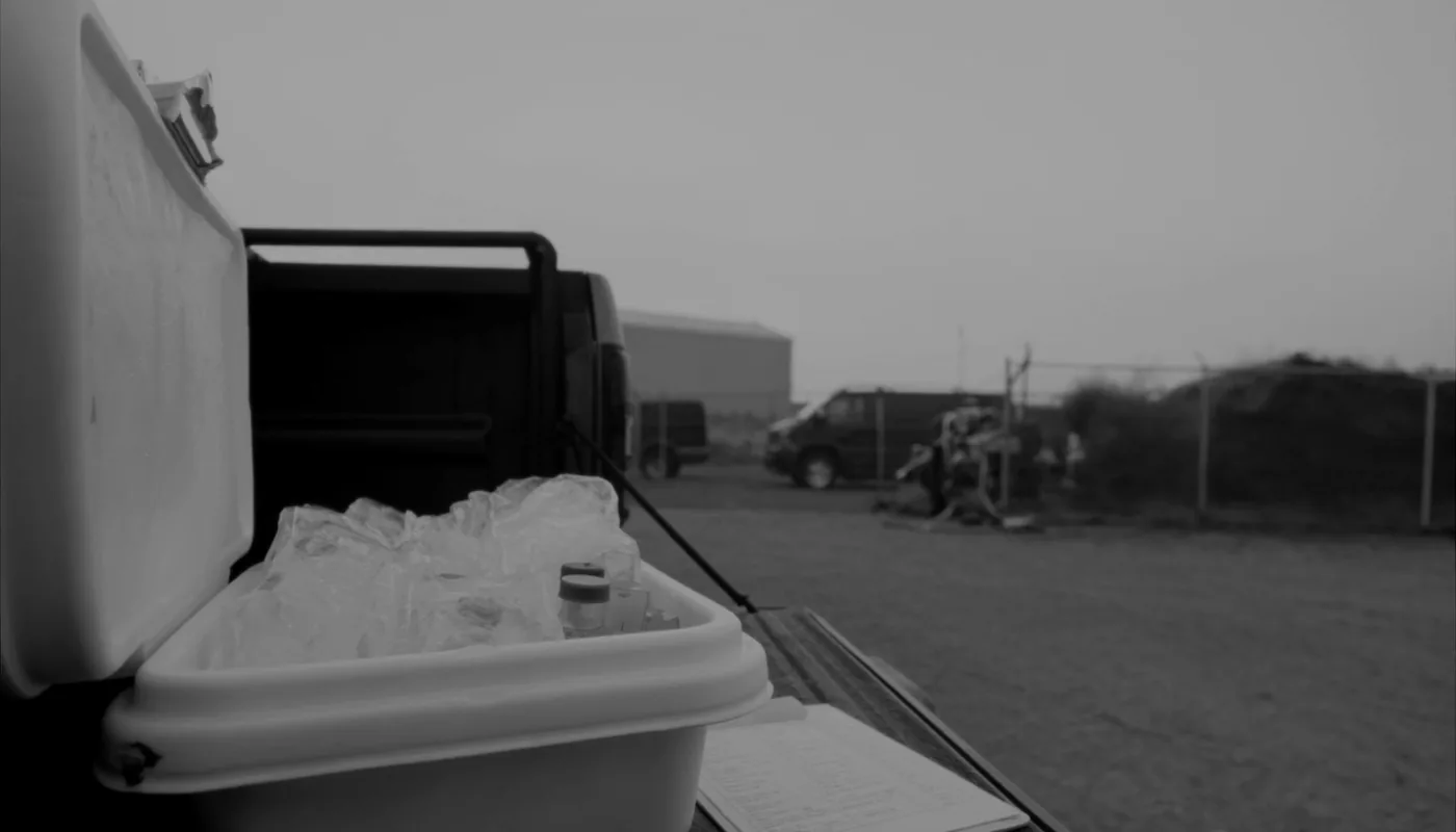 An open hard sided sample cooler on a pickup tailgate, packed with bagged ice and upright plastic sample bottles, with a clipboard beside it.